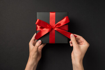 First person top view photo of hands unpacking black giftbox with red ribbon bow on isolated black background