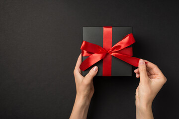 First person top view photo of girl hands unpacking black giftbox with vivid red ribbon bow on isolated black background with copyspace