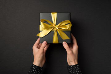 First person top view photo of hands in polka dot shirt giving black giftbox with golden satin ribbon bow on isolated black background