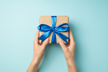 First person top view photo of hands holding craft paper giftbox with blue ribbon bow on isolated pastel blue background