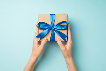 First person top view photo of girl's hands holding craft paper giftbox with blue satin ribbon bow on isolated pastel blue background