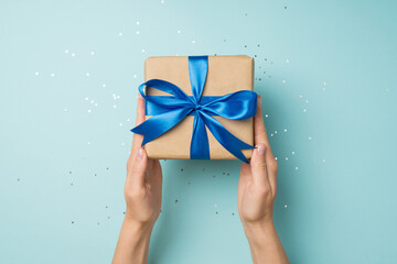 First person top view photo of woman's hands holding craft paper giftbox with blue ribbon bow over shiny confetti on isolated pastel blue background