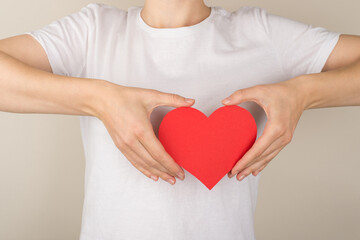 Cropped closeup photo of young woman in white t-shirt holding red paper heart near breast on isolated grey background