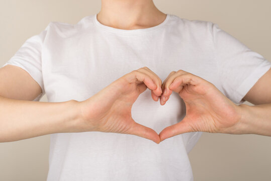 Cropped closeup photo of girl in white t-shirt making heart with fingers near left side of chest on isolated grey background