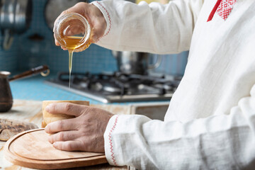 Ethnic linen shirt on the man. A man with a beard in the kitchen pours honey into a cup. Cooking dessert in a rustic kitchen.