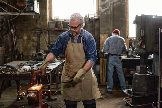 Mature bearded blacksmith in apron working with iron in the workshop with his colleague in the background