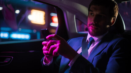 Successful Businessman in a Suit Commuting from Office in a Backseat of His Car at Night. Entrepreneur Using Smartphone while in Transfer Taxi in Urban City Street with Working Neon Signs.