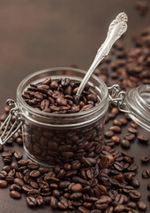 Roasted coffee beans in glass jar with steel spoon on brown background.