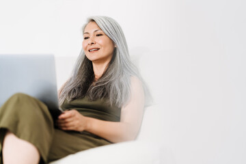 Naklejka premium Mature asian woman working with laptop while sitting on couch