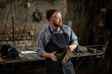 Mature blacksmith in apron holding protective gloves standing near his workplace in the workshop