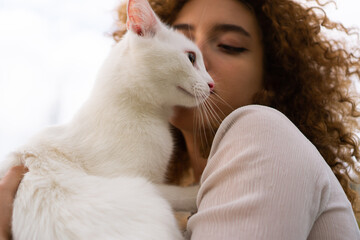 A beautiful teenage girl with curly hair holds a white cat close to her face. She is enjoying time with her pet.