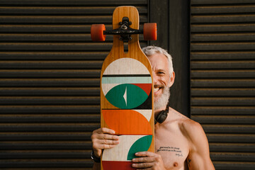 Shirtless mature man smiling while standing with skateboard