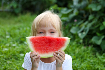 Blonde child with a piece of watermelon in his hands sitting in the garden on the green grass. A piece of watermelon instead of a smile