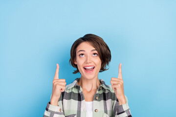 Photo of cute impressed young lady wear plaid shirt pointing fingers up looking empty space isolated blue color background