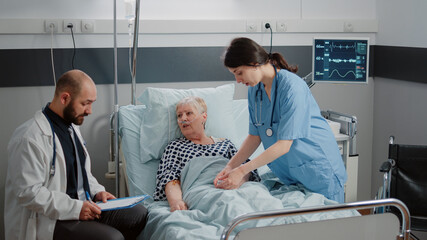 Doctor and nurse doing consultation with retired patient in hospital ward. Medical team examining diagnosis and treatment progress for ill woman with oximeter and nasal oxygen tube