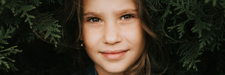 Obraz premium portrait of the face of a cute happy caucasian candid healthy eight year old kid girl surrounded by branches and leaves of green plant thuja or cypress on nature outdoor. banner