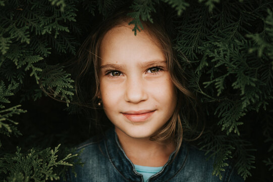 portrait of the face of a cute happy caucasian candid healthy eight year old kid girl surrounded by branches and leaves of green plant thuja or cypress on nature outdoor