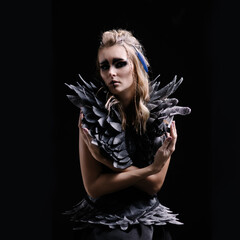 Beautiful woman in makeup and costume for halloween on a black background. Fantasy image of a harpy or valkyrie. Celtic mythology character. Wings and feathers. Smokey eyes. Carnival girl black bird - obrazy, fototapety, plakaty