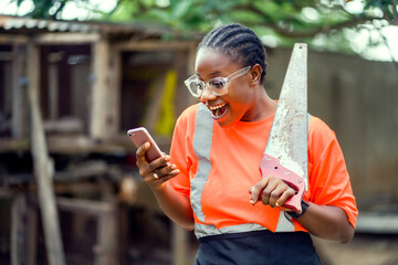 image of excited african lady with smart phone and a working tool on her shoulder