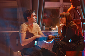 Young musician with guitar talking to young woman while they sitting at a party