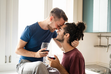 Tenderness moment of smiling gay couple, young men toasting with red wine in the kitchen, male...