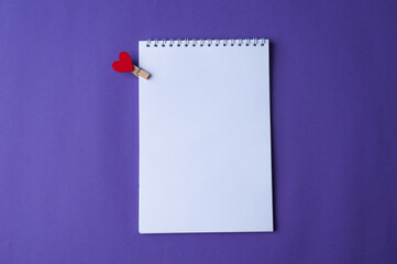 A notepad to write a love note for the loved one, with heart pin on purple background. Top view. Copy space.