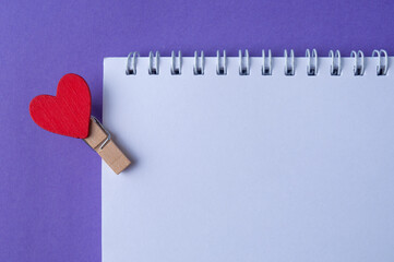 A notepad to write a love note for the loved one, with heart pin on purple background. Top view. Copy space.