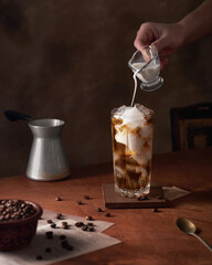 woman pours cream into a glass of cold coffee, dark background