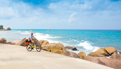 Plakat Happy young guy riding a bike on the road by the sea in the Seychelles. Concept of vacation and active lifestyle. Cycling. The man on the bike.
