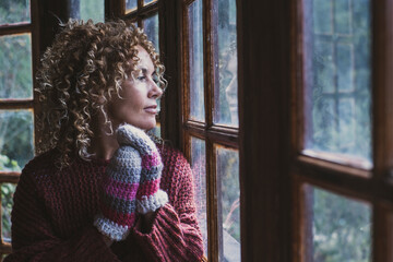 Adult pretty woman looking outside the window at home in winter season. Thoughtful happy female people enjoy cold season using gloves at home and dreaming. Romantic portrait of lady