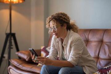 Serious young woman text messaging using mobile phone while sitting on sofa at home. Woman with curly hair and eyeglasses reading or texting using mobile phone in the living room - Powered by Adobe