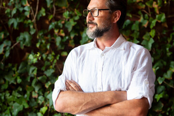 Thoughtful mature man wearing eyeglasses and looking away while standing with his arms crossed in front of green leaves background. Bearded man looking to his side in front of ivy creeper background