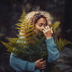Happy young woman in blue sweater holding fresh fragile fern leaves plant in forest or park....