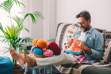 Mature man knitting wool with needle while sitting on sofa at home. Caucasian man smiling and making sweater of wool while relaxing at home. Woollen balls in basket on table.