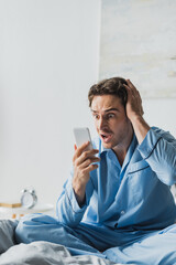 Scared man in pajama looking at smartphone on bed in morning