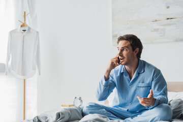 Stressed man in pajama talking on cellphone on bed in morning