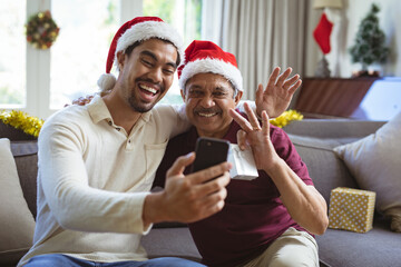 Happy biracial adult son and senior father in santa hats making smartphone christmas video call - Powered by Adobe