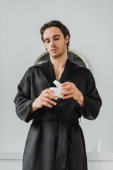 Young man in satin robe holding blurred jar with cosmetic cream in bathroom