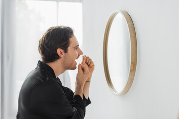 Side view of man in satin robe using dental floss near mirror in bathroom