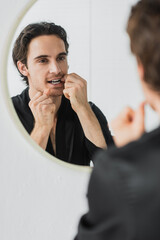Blurred man in robe looking at mirror while cleaning teeth with dental floss