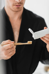 Cropped view of blurred man in black robe holding toothbrush and toothpaste in bathroom