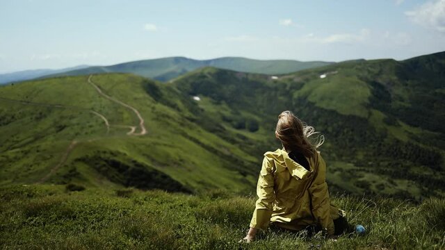 Beautiful blonde woman tourist in a bright yellow jacket sits on top of mountain and stretching out hands looking at Mountain valley. Gold hair in fresh air wind