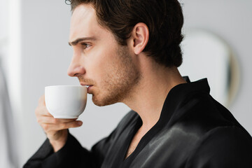 Side view of young man in robe holding coffee in bathroom