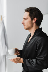 Side view of man in silk robe holding cup of coffee in bathroom