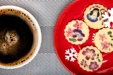 Beautiful edible flower cookies with Christmas ornament snowflakes on a bright red plate with a cup of black coffee. - Powered by Adobe