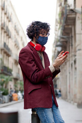 Young Caucasian man with glasses, mask and red headset texting from his cell phone outdoors. Red mobil cover. Photograph taken from the side. Lifestyle concept.