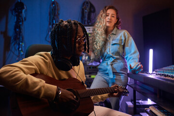 Fototapeta na wymiar African young musician playing guitar while young woman singer in the background in the recording studio