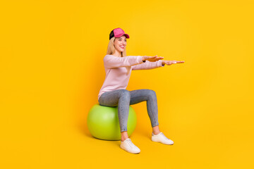 Photo of funky pretty young woman dressed pink pullover headwear sitting fitball looking empty space isolated yellow color background