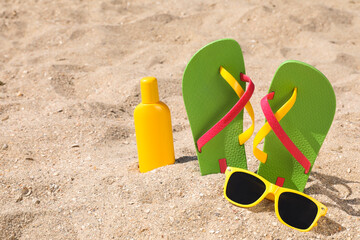 Stylish flip flops, sunglasses and sun protection cream on sand, space for text