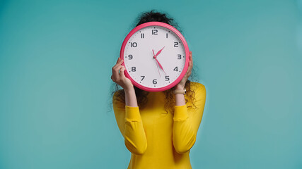 young woman obscuring face with wall clock isolated on blue